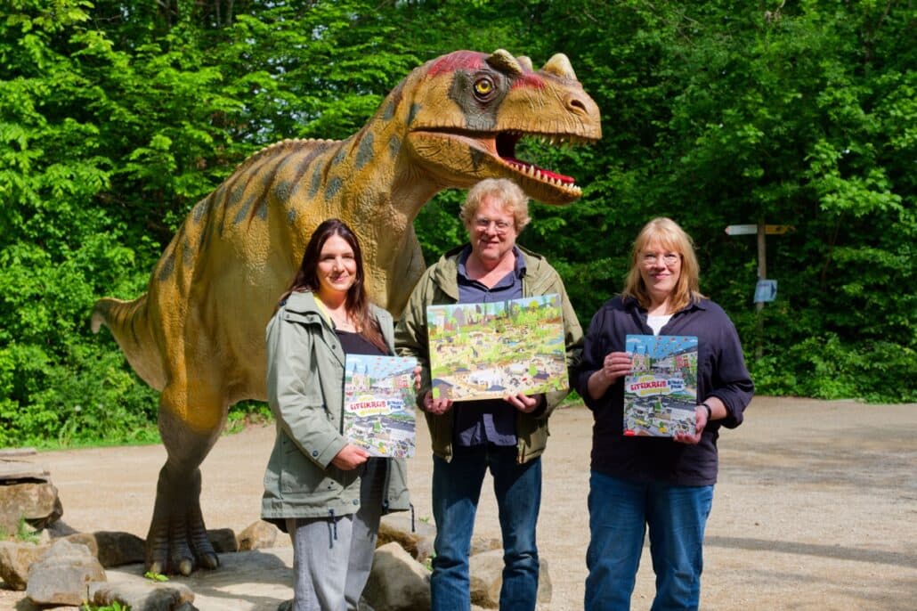 Eifelkreis Wimmelbuch Vorstellung mit Lorette Englich, Bruno Zwank und Elke Wagner im Dinosaurierpark Teufelsschlucht. Drei Personen mit Dinosauriermodell.